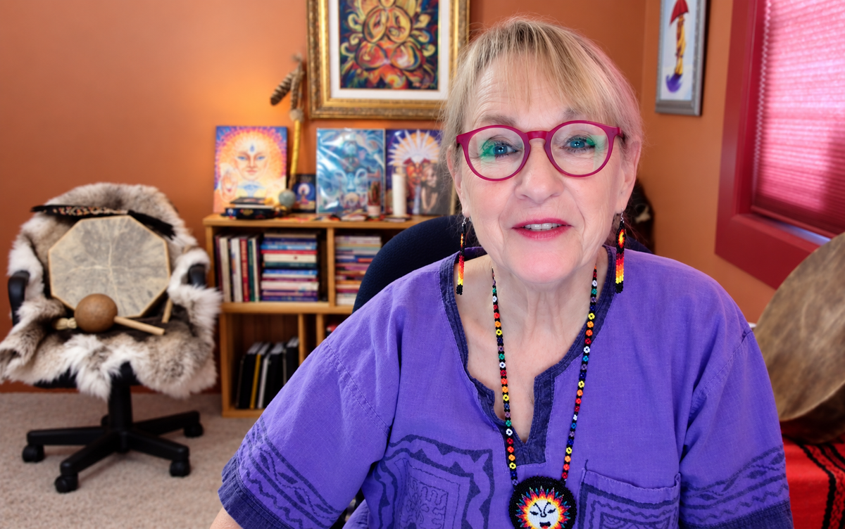 Katherine Skaggs in shamanic shirt and attire, sitting in a office with shelves, framed art, and musical instruments in the background.