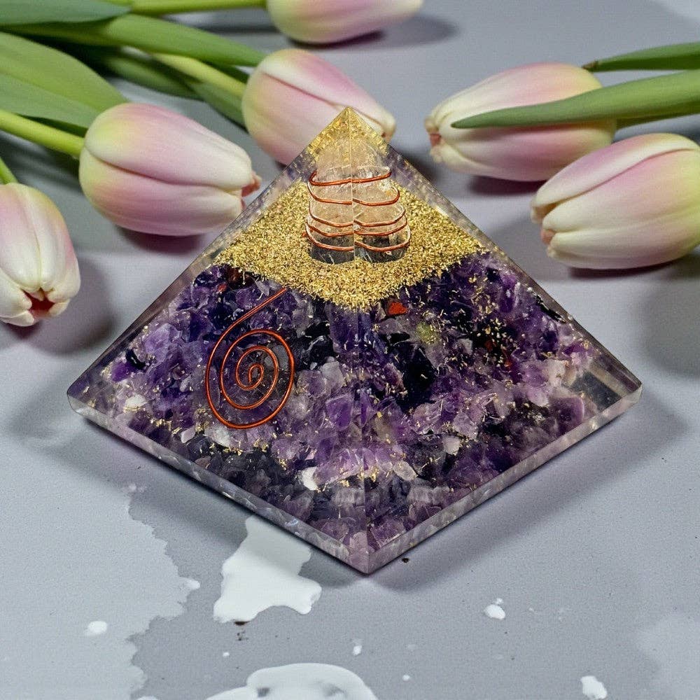 Decorative pyramid with purple amethyst crystal, copper spiral, copper wrap quartz, and gold and copper shavings sitting on a gray surface with pink tulips.