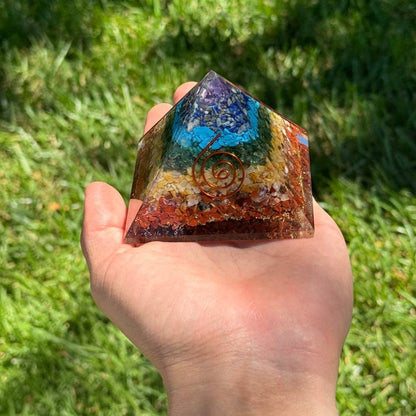 Hand holding a colorful organite chakra crystal pyramid with a spiral design against a grassy background