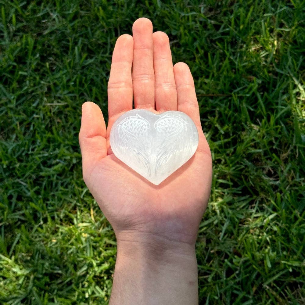 Selenite heart with angel wings held in palm of hand grass background