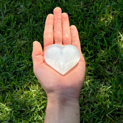 Selenite heart with angel wings held in palm of hand grass background