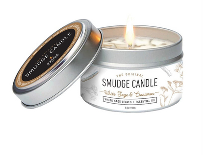 An open silver tin candle labeled White Sage & Cinnamon 'SMUDGE CANDLE' with a lit wick, shown beside its matching lid on a clean white background. The label reads 'White Sage & Cinnamon' and 'White Sage Leaves + Essential Oil.